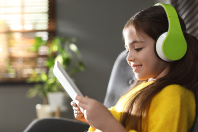 Photo of Cute little girl with headphones and tablet listening to audiobook at home