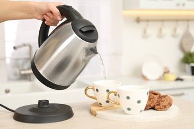 Photo of Woman pouring hot water from electric kettle into cup in kitchen, closeup. Space for text