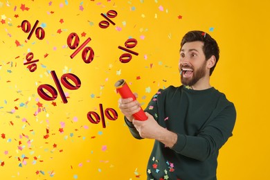 Image of Discount offer. Happy man blowing up party popper on golden background. Confetti and percent signs in air