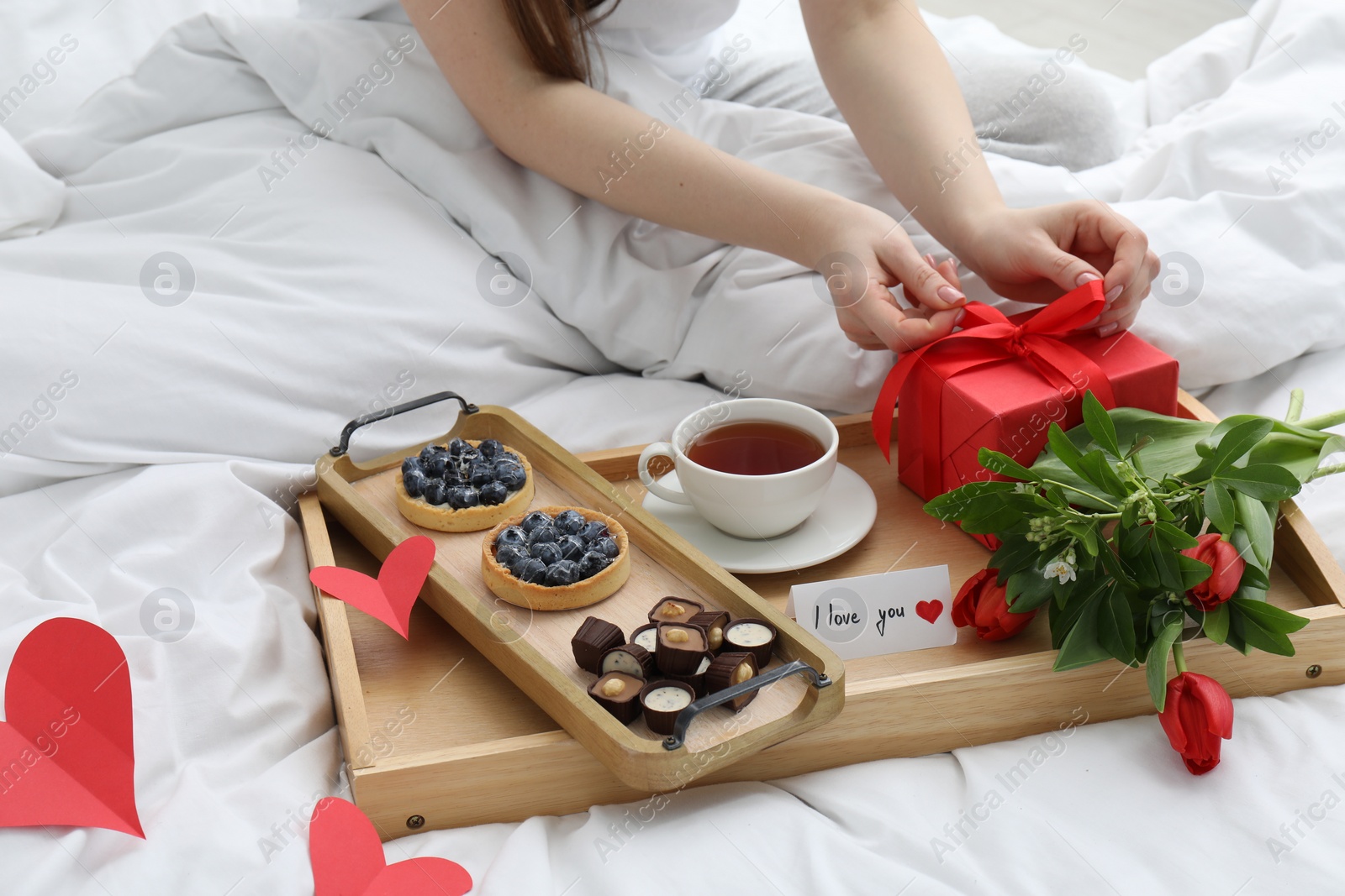 Photo of Tasty breakfast served in bed. Woman with gift box, tea, desserts, flowers and I Love You card at home, closeup