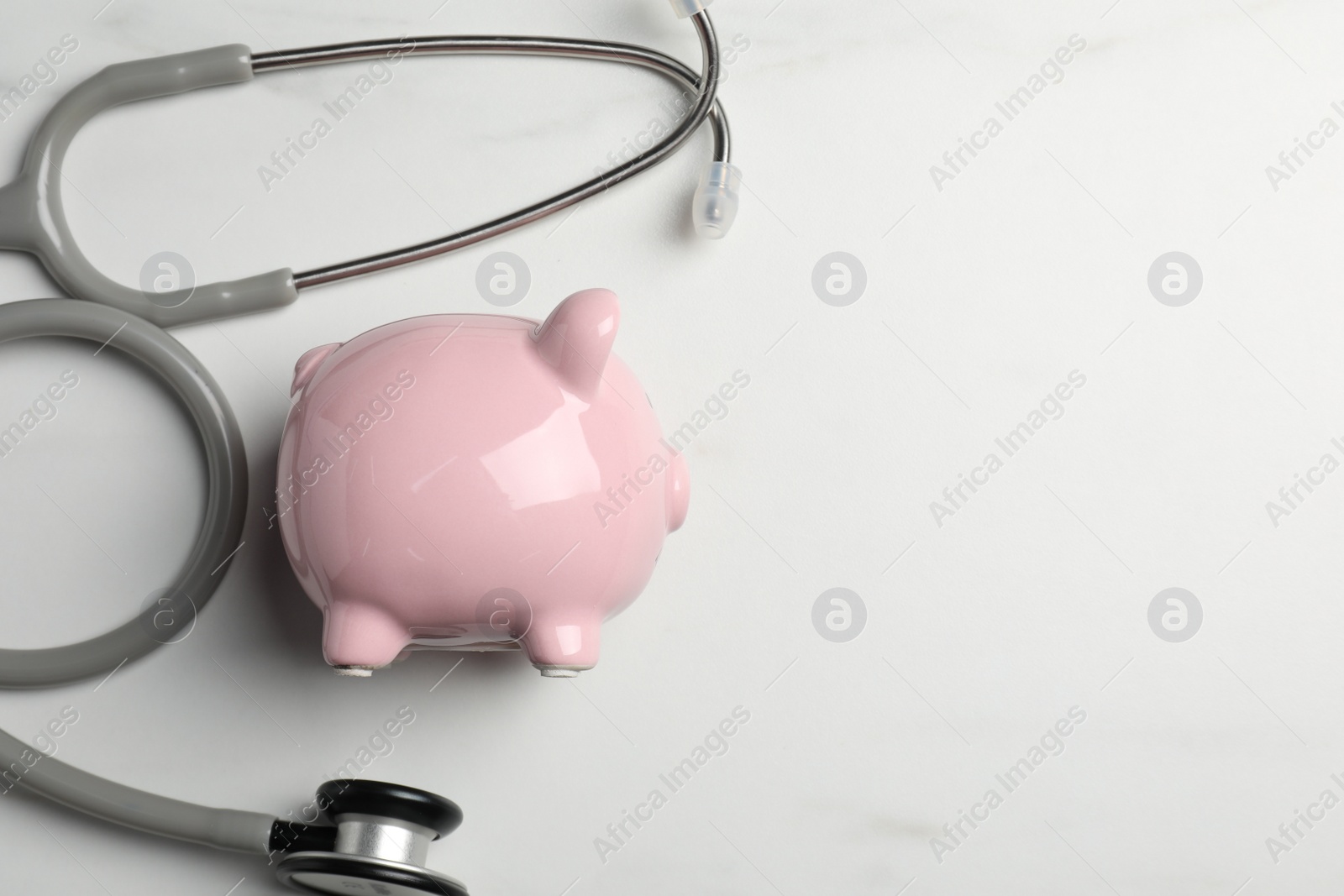 Photo of Pale pink ceramic piggy bank and stethoscope on white table, flat lay with space for text. Medical insurance