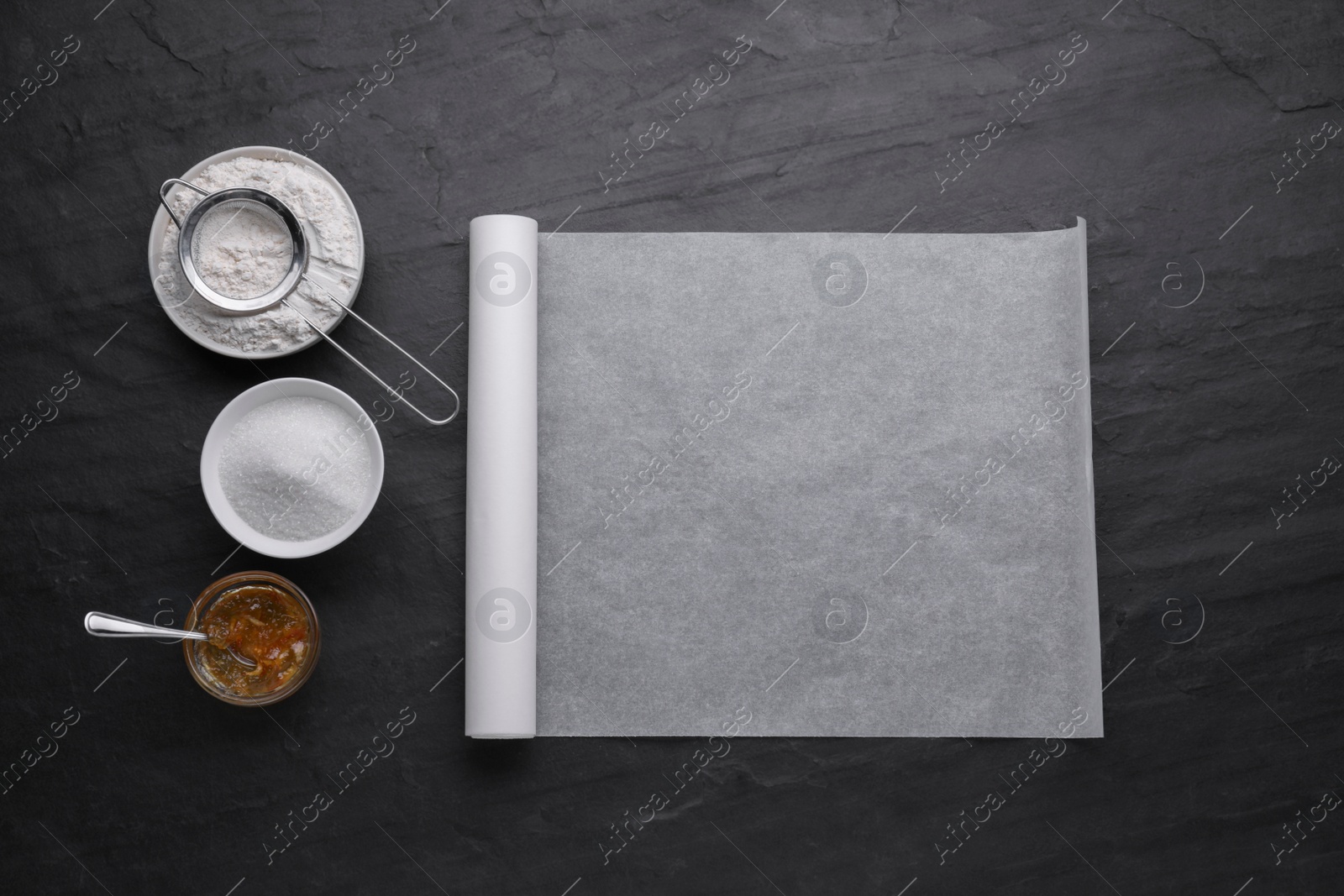 Photo of Roll of baking parchment paper and ingredients on black slate background, flat lay