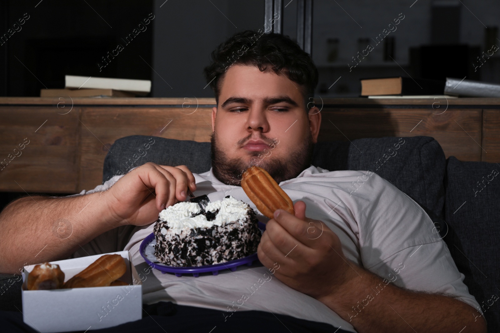 Photo of Depressed overweight man eating sweets in living room at night