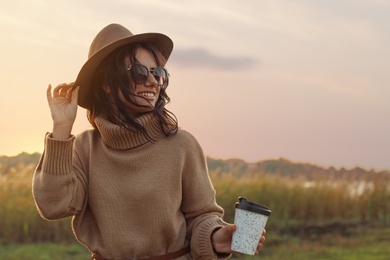 Beautiful young woman with cup of coffee wearing stylish autumn sweater outdoors