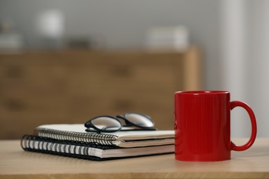 Red ceramic mug, notebooks and glasses on wooden table at workplace. Space for text