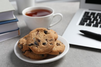 Photo of Chocolate chip cookies, cup of tea and laptop on light grey table in office, closeup