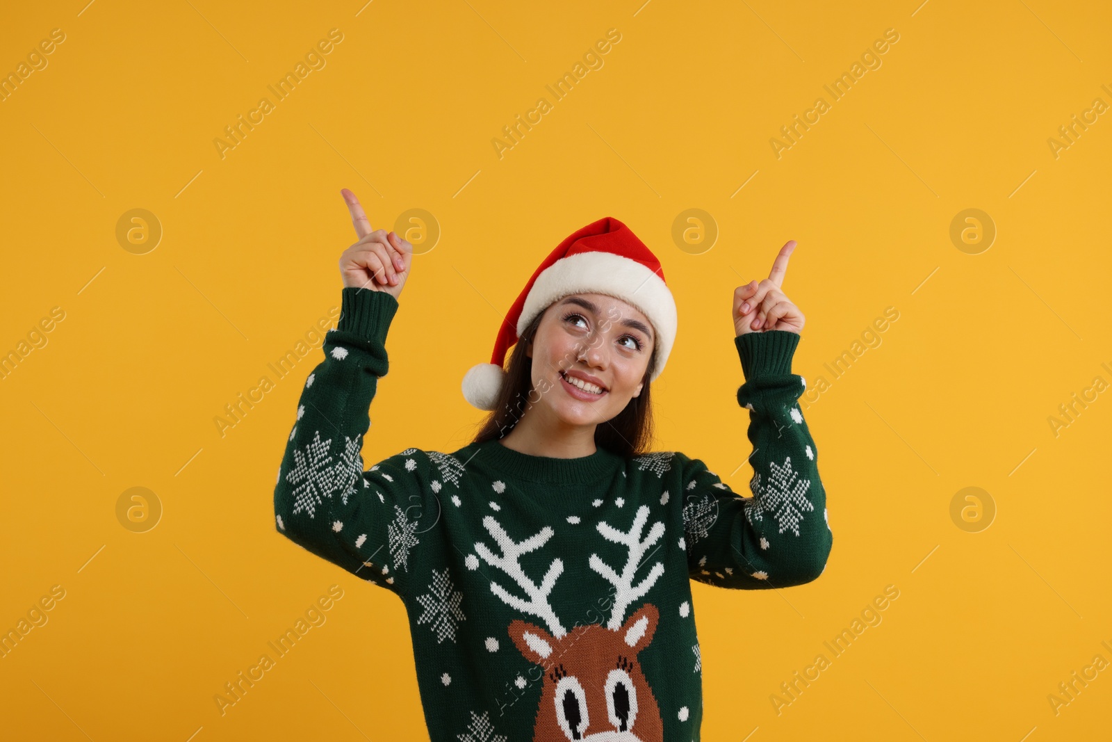 Photo of Happy young woman in Christmas sweater and Santa hat pointing at something on orange background