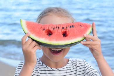 Photo of Cute little girl holding slice of watermelon with holes in front of her face on beach