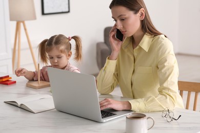 Photo of Woman working remotely at home. Mother using laptop while daughter playing at desk