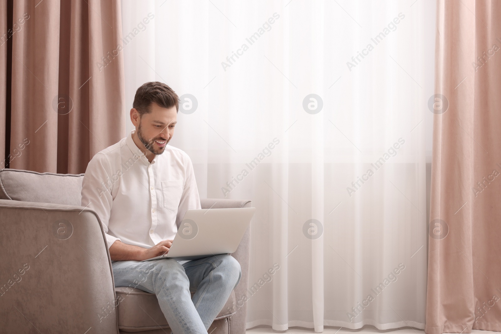 Photo of Smiling man holding laptop on armchair near window with beautiful curtains at home. Space for text