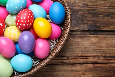 Photo of Colorful Easter eggs in wicker basket on wooden table, top view. Space for text