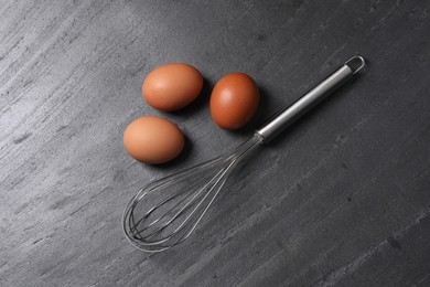 Metal whisk and raw eggs on dark grey table, flat lay