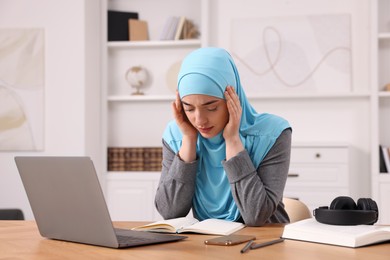 Tired Muslim woman working near laptop at wooden table in room
