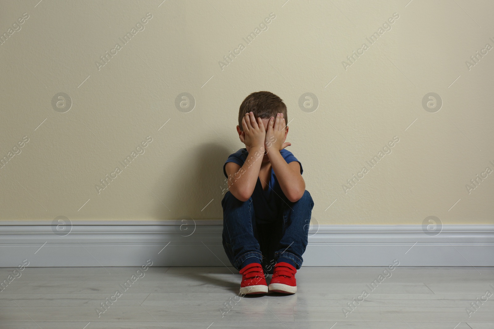 Photo of Little boy closing face with hands on floor near yellow wall. Child in danger