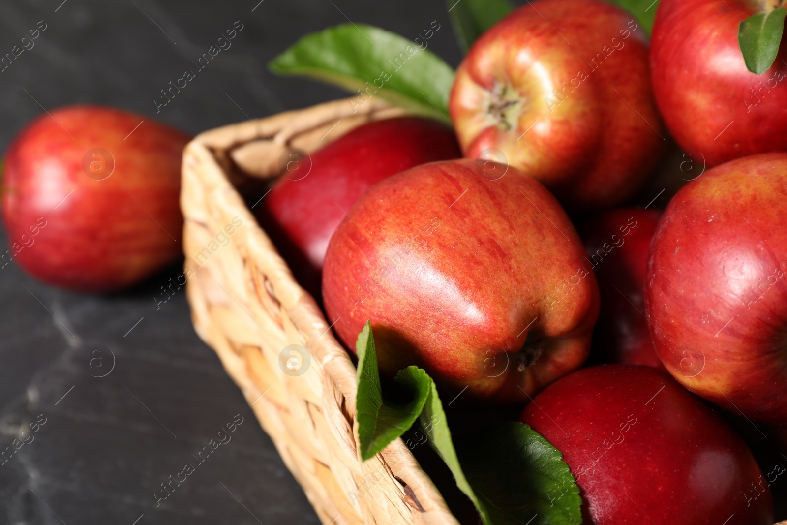 Photo of Fresh red apples and leaves in basket on dark grey table, closeup