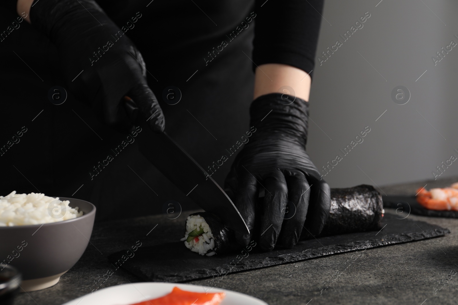 Photo of Chef in gloves cutting sushi roll at dark textured table, closeup