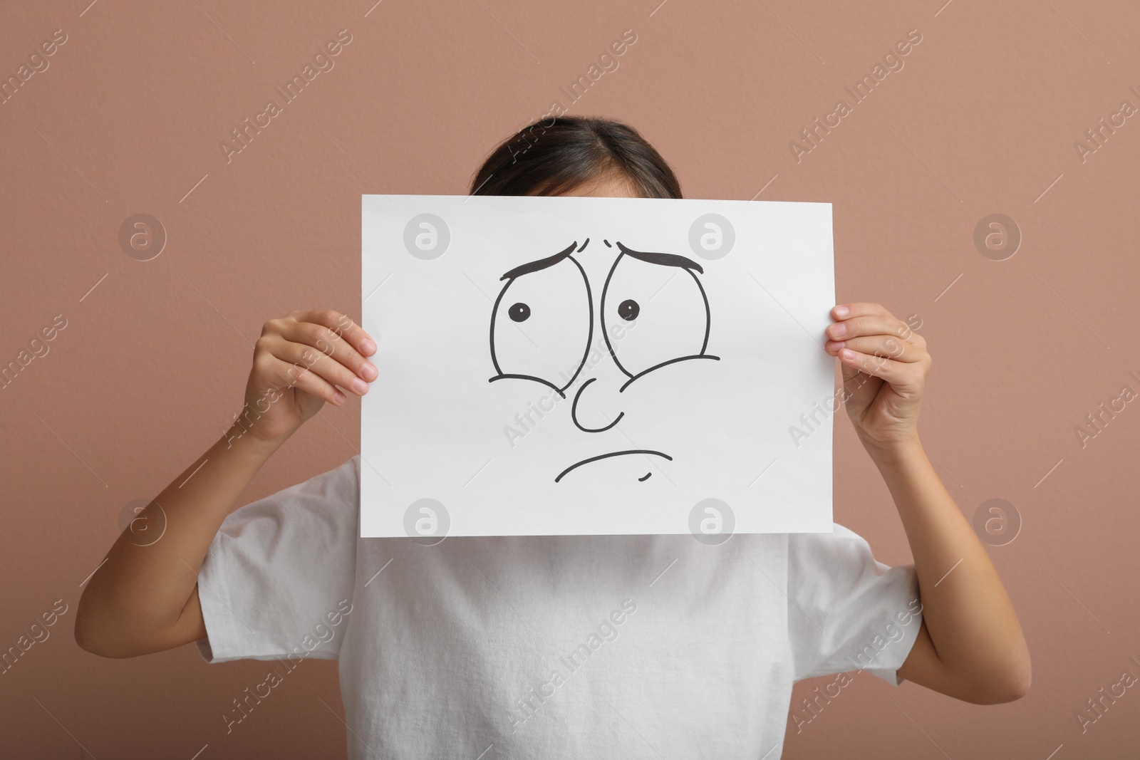 Photo of Little girl hiding behind sheet of paper with sad face on pale pink background