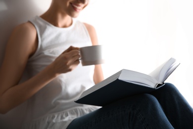 Photo of Young woman with cup of coffee reading book on light background, closeup