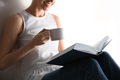 Young woman with cup of coffee reading book on light background, closeup