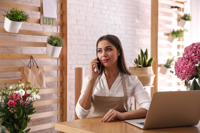 Photo of Florist talking on smartphone near laptop in workshop