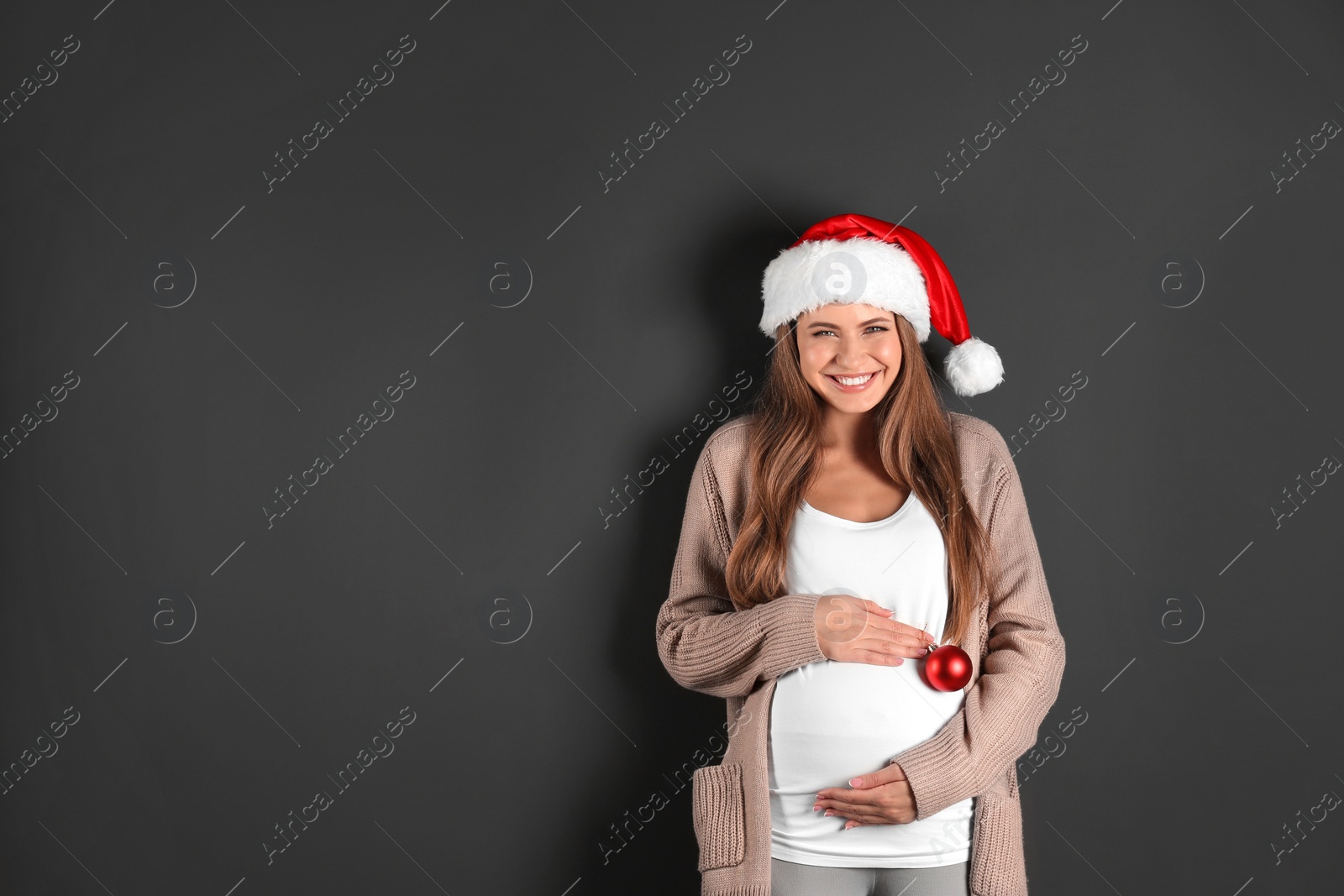 Photo of Young pregnant woman in Santa hat with Christmas decoration on grey background, space for text