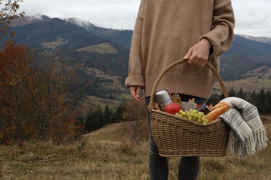 Woman holding wicker picnic basket with thermos, snacks and plaid in mountains on autumn day, closeup. Space for text