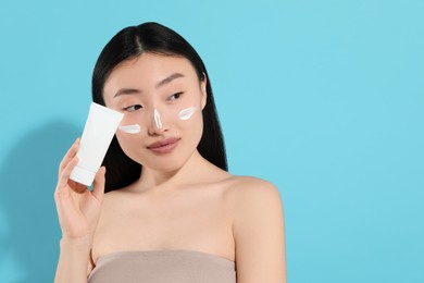 Beautiful young woman with sunscreen on her face holding sun protection cream against light blue background, space for text