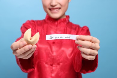 Photo of Young woman holding tasty fortune cookie with prediction You Can Do This on light blue background, closeup