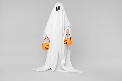 Child in white ghost costume holding pumpkin buckets on light grey background. Halloween celebration