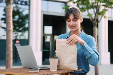 Photo of Happy businesswoman with paper bag having lunch at wooden table outdoors
