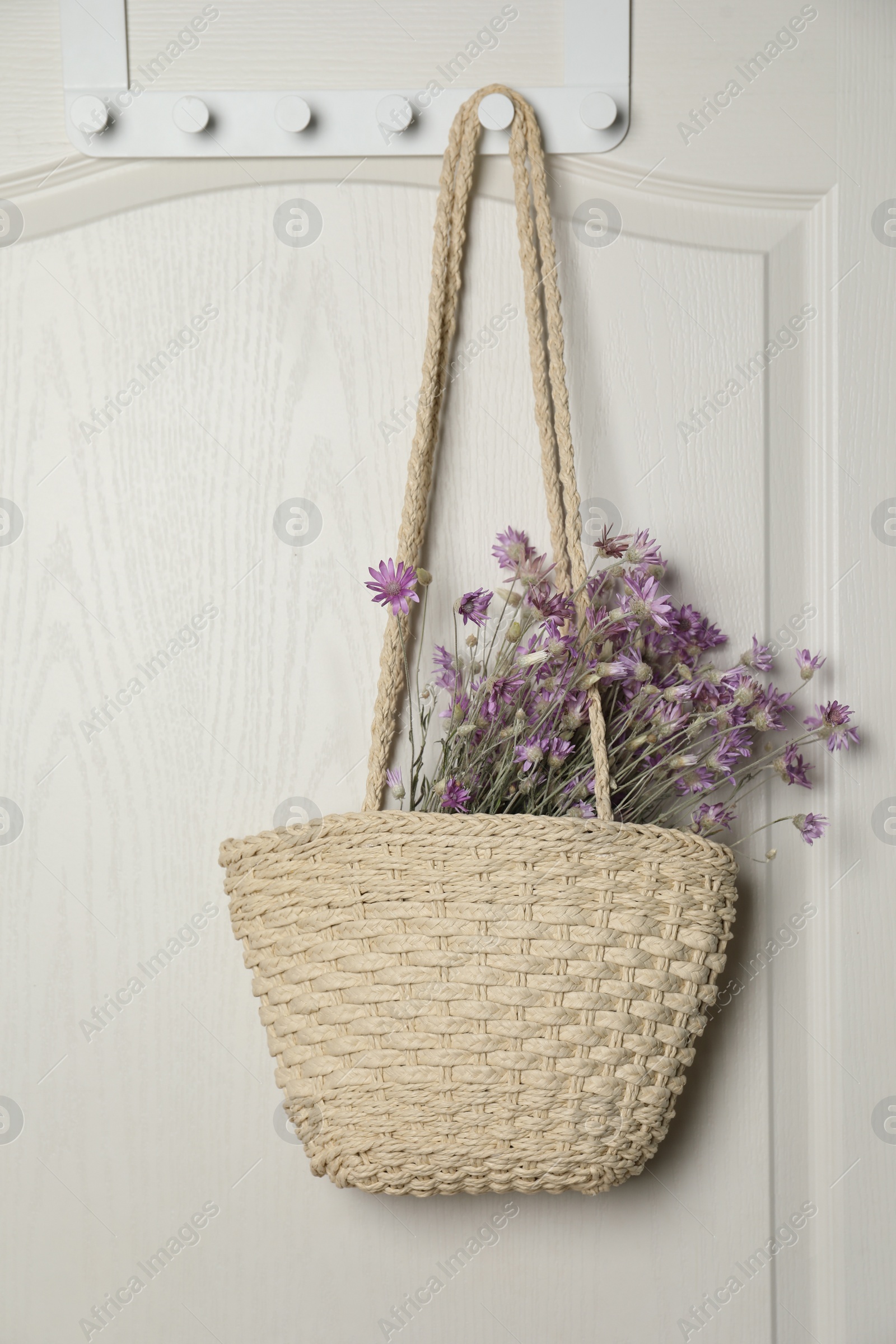 Photo of Stylish beach bag with beautiful bouquet of wildflowers hanging on white wooden door