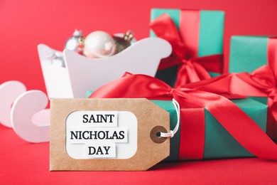 Photo of Gift box and tag with text Saint Nicholas Day on red background, closeup