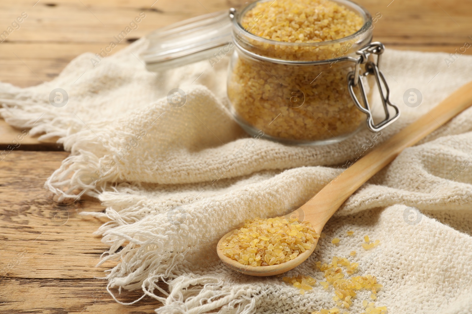 Photo of Glass jar and spoon with raw bulgur on table, closeup