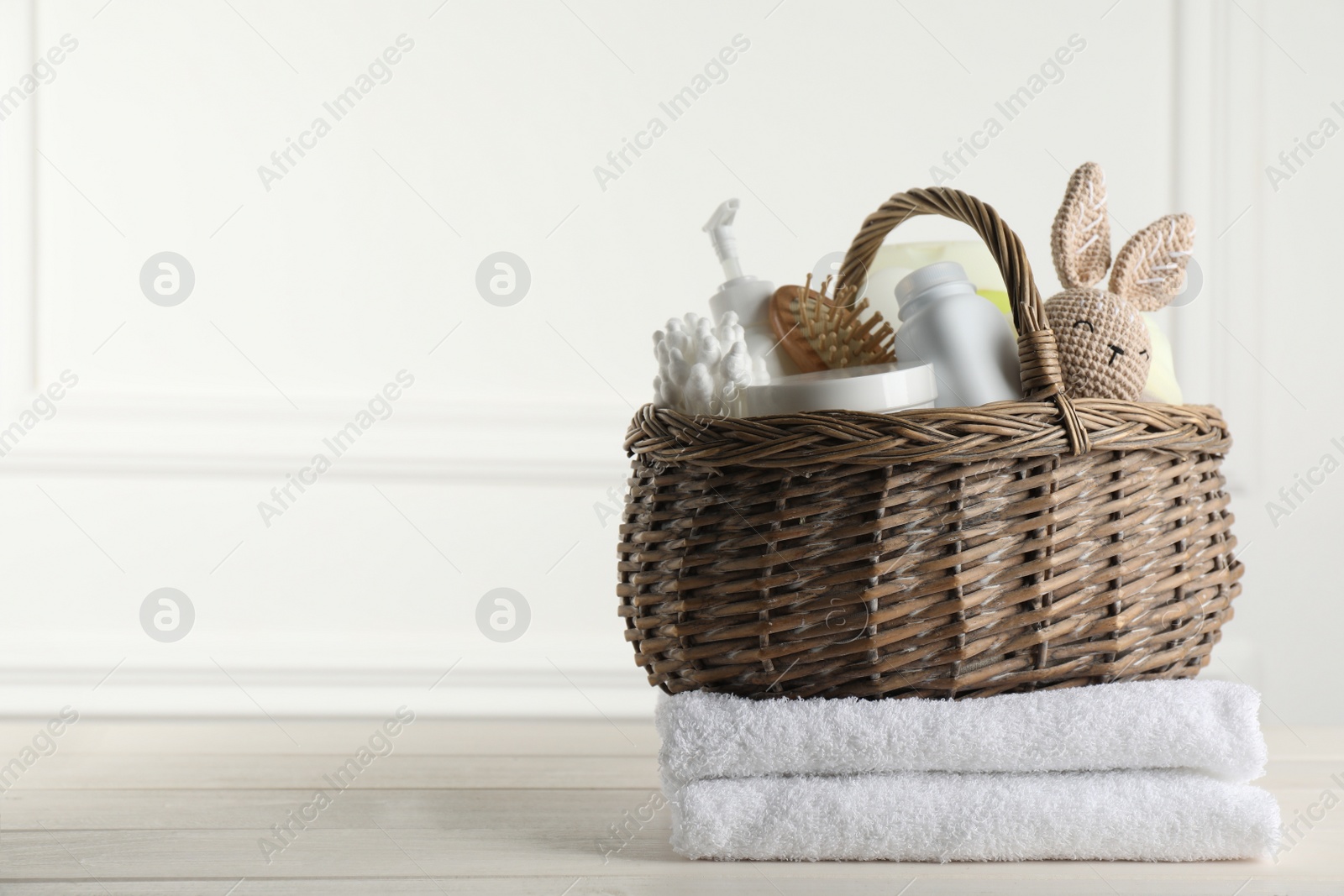 Photo of Wicker basket with different baby cosmetic products, accessories and toy on white wooden table. Space for text