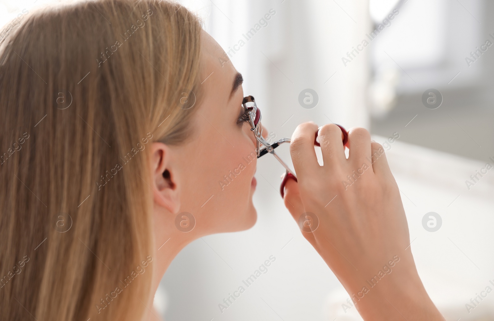 Photo of Attractive young woman curling her eyelashes indoors