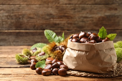 Photo of Fresh sweet edible chestnuts and paper bag on wooden table. Space for text