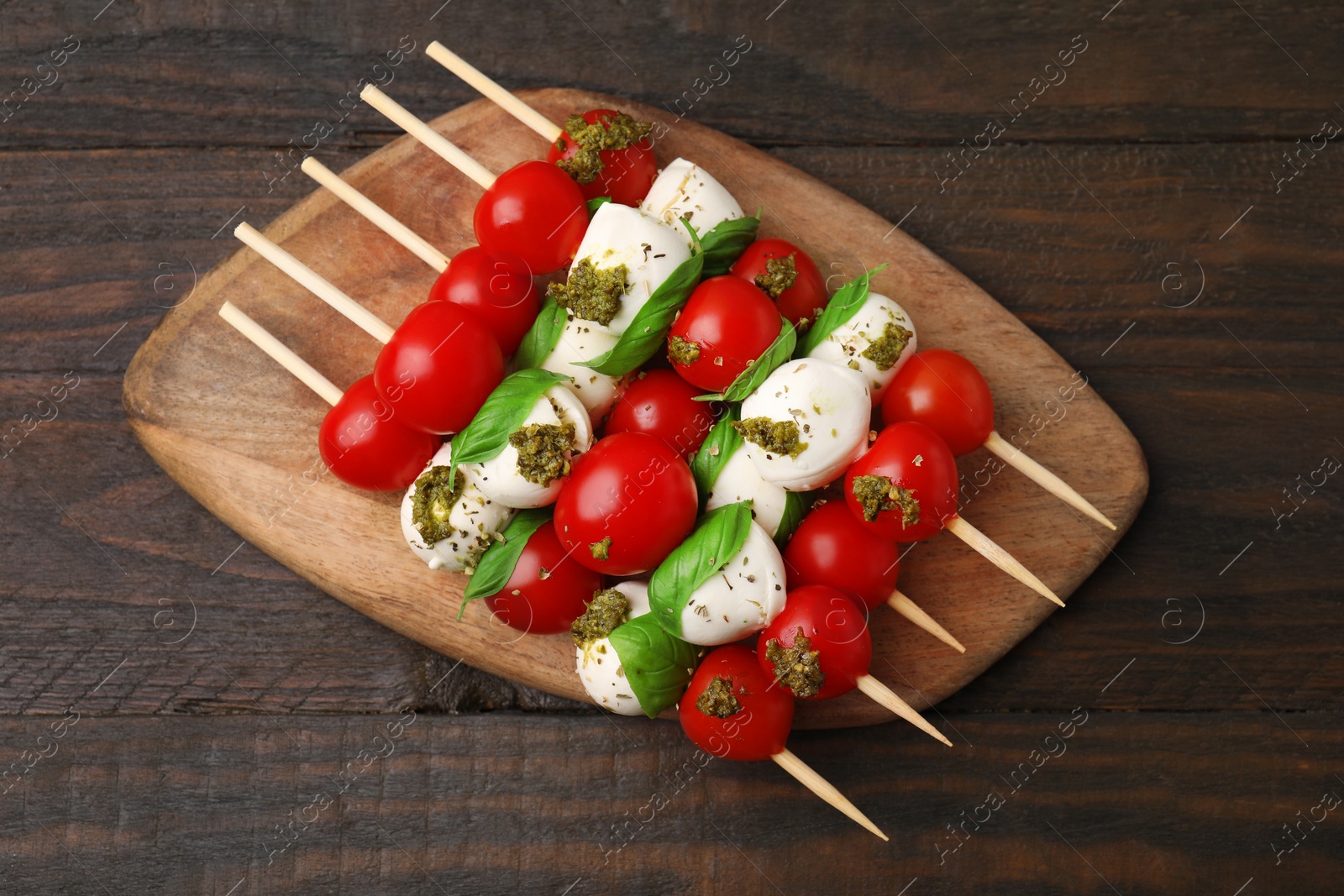 Photo of Caprese skewers with tomatoes, mozzarella balls, basil and pesto sauce on wooden table, top view