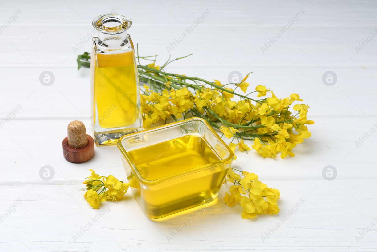 Photo of Rapeseed oil in glass bowl, bottle and beautiful yellow flowers on white wooden table