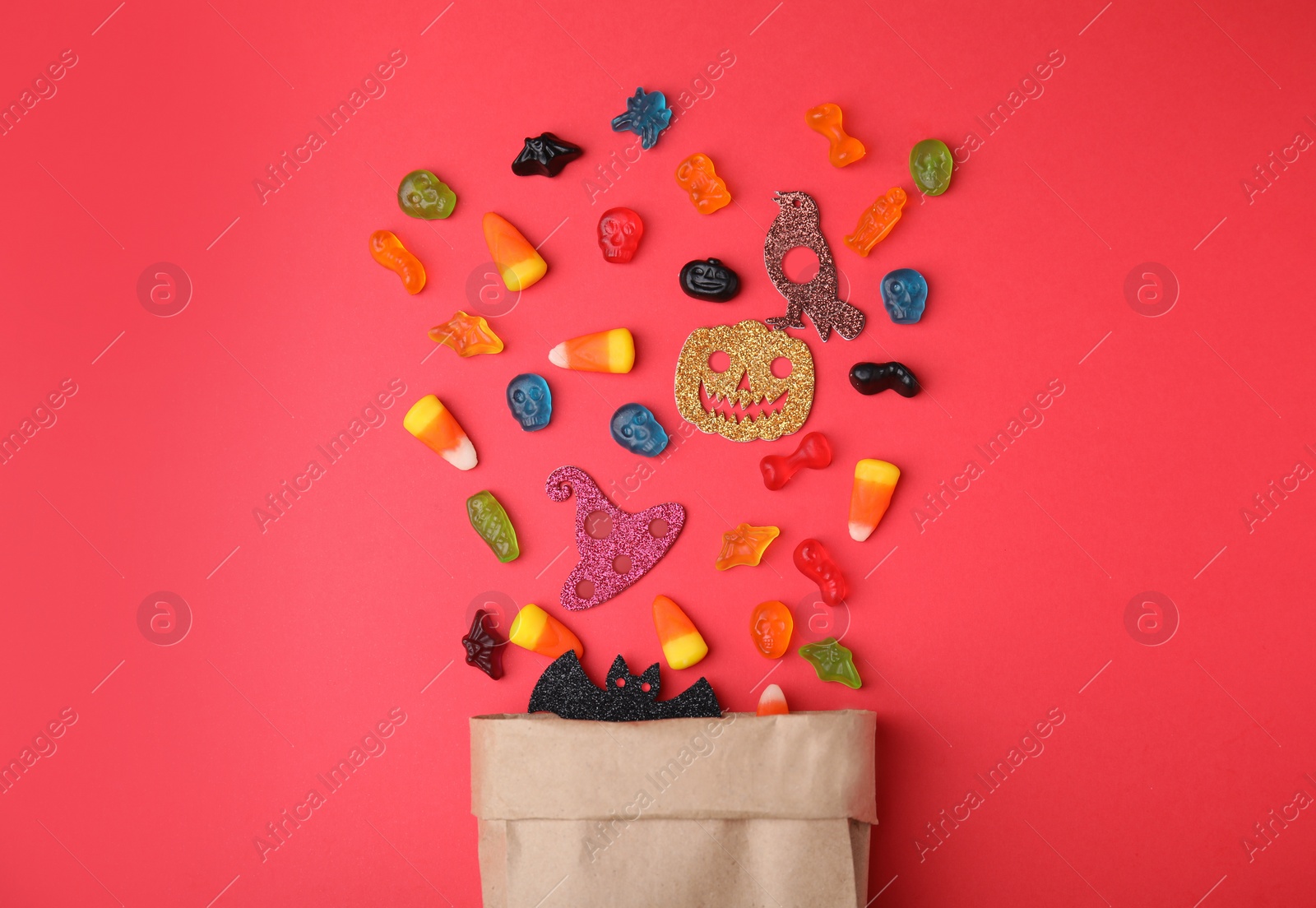 Photo of Paper bag of tasty candies and Halloween decorations on red background, flat lay