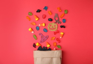 Paper bag of tasty candies and Halloween decorations on red background, flat lay