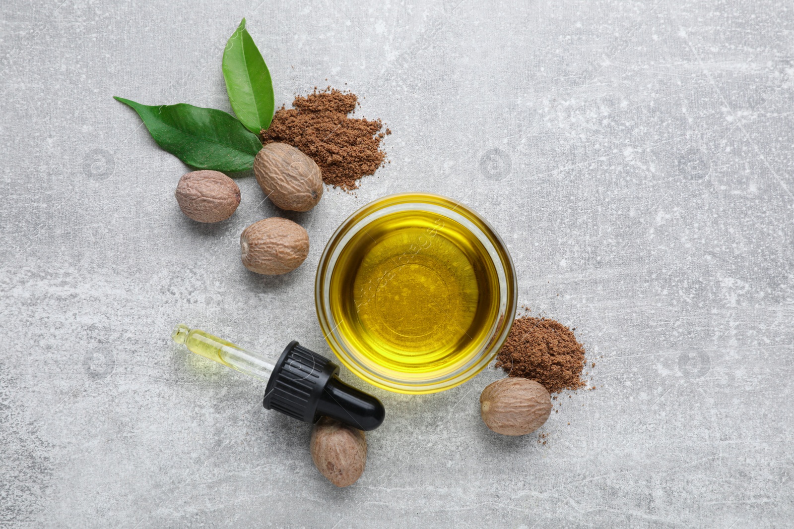 Photo of Bowl of nutmeg oil, pipette, nuts and powder on light grey table, flat lay