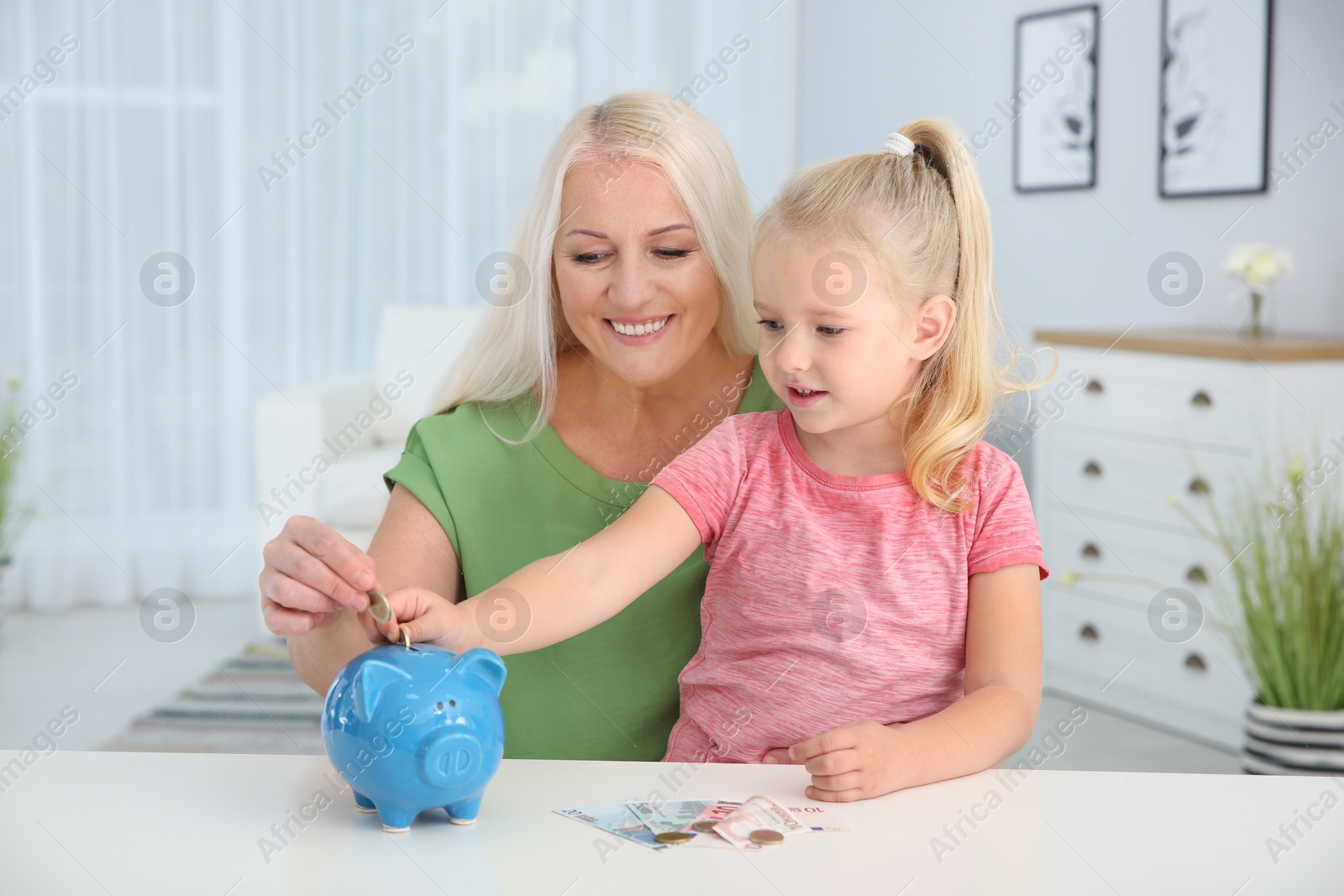 Photo of Little girl with grandmother putting money into piggy bank at table