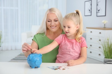 Photo of Little girl with grandmother putting money into piggy bank at table