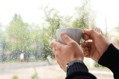 Photo of Young man with cup of coffee near window indoors on rainy day, closeup