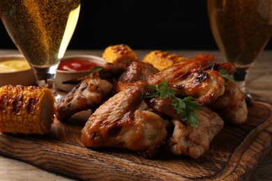 Delicious baked chicken wings, grilled corn and glasses with beer on wooden table, closeup