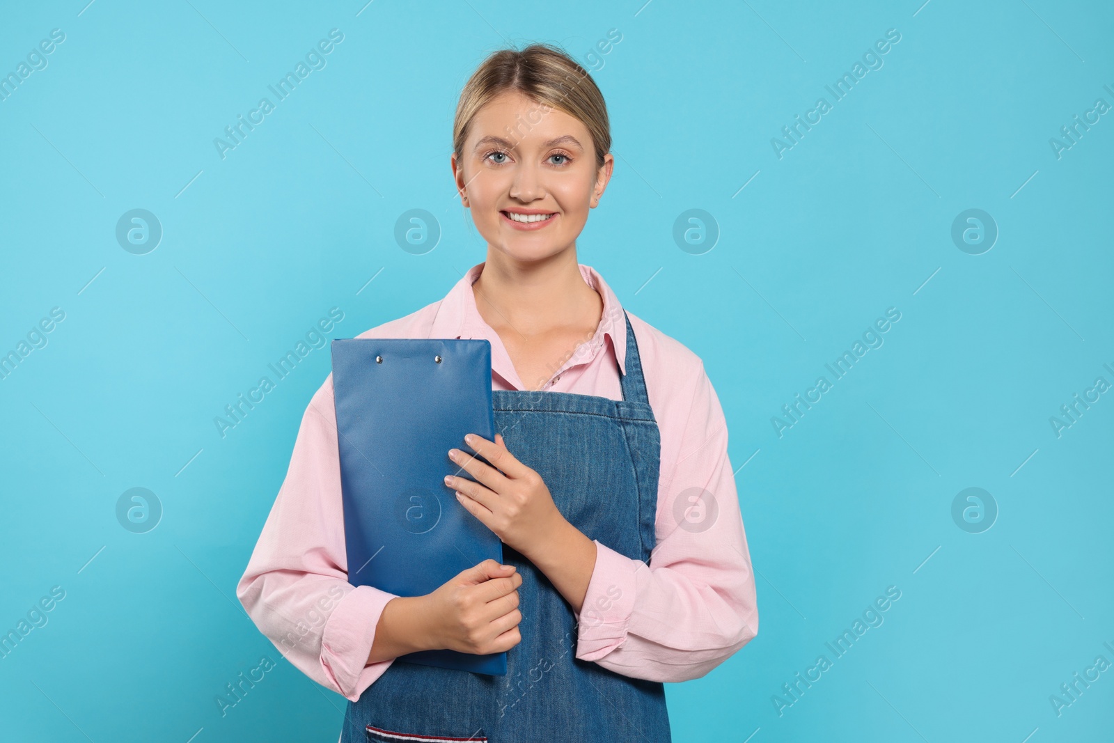 Photo of Beautiful young woman in denim apron with clipboard on light blue background