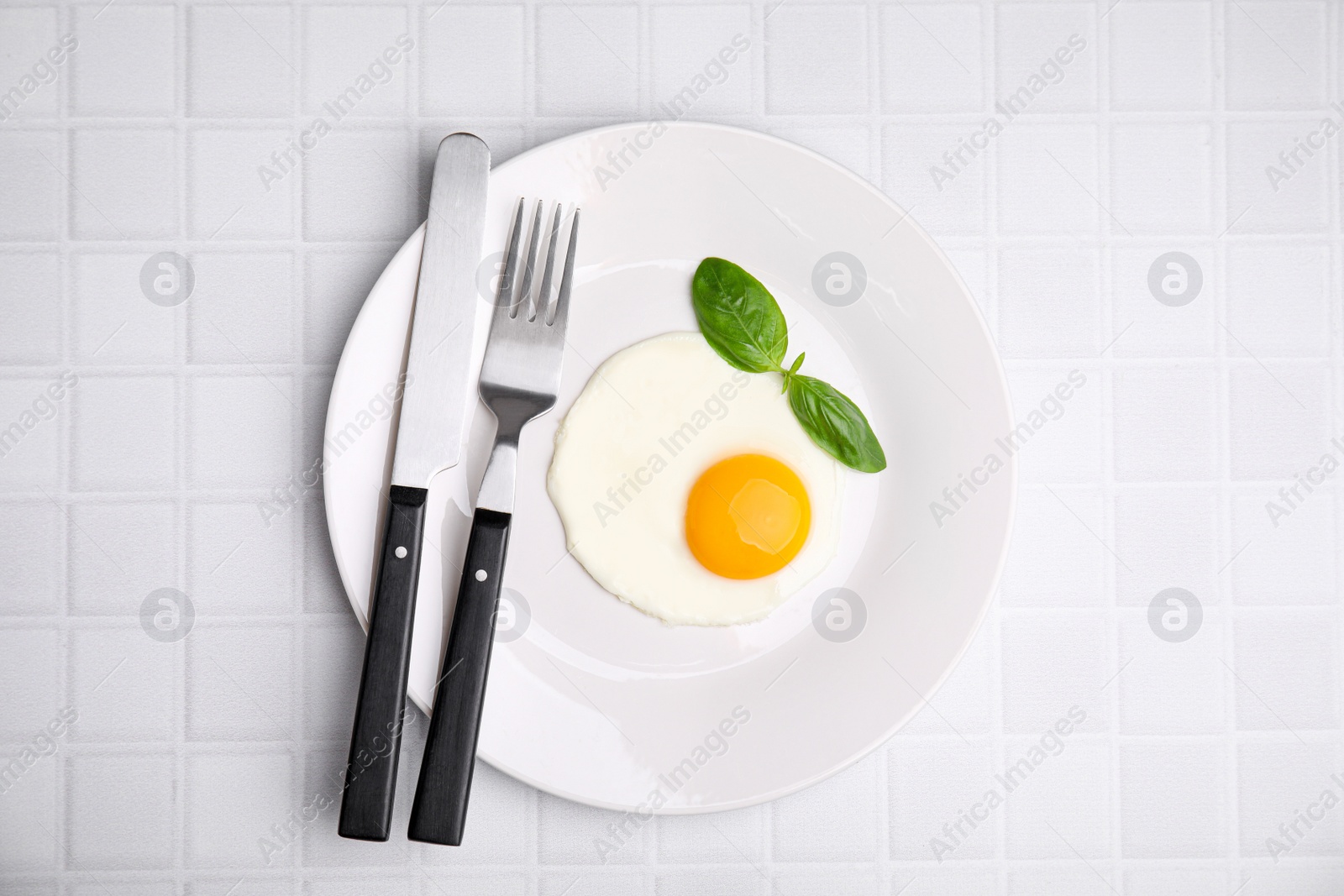 Photo of Tasty fried egg with basil in plate on white tiled table, top view