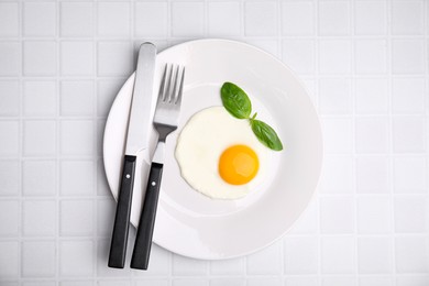 Tasty fried egg with basil in plate on white tiled table, top view