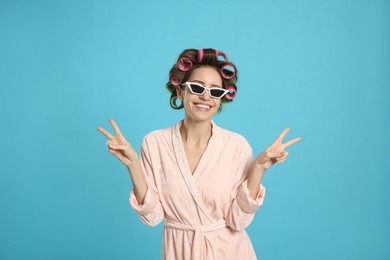 Beautiful young woman in bathrobe with hair curlers and sunglasses on light blue background