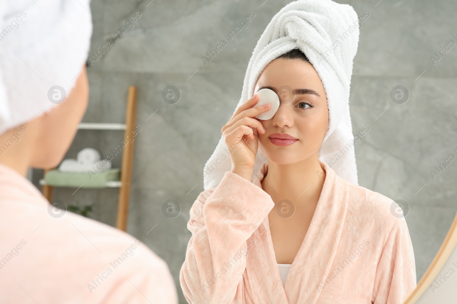 Photo of Beautiful woman in terry towel removing makeup with cotton pad near mirror indoors